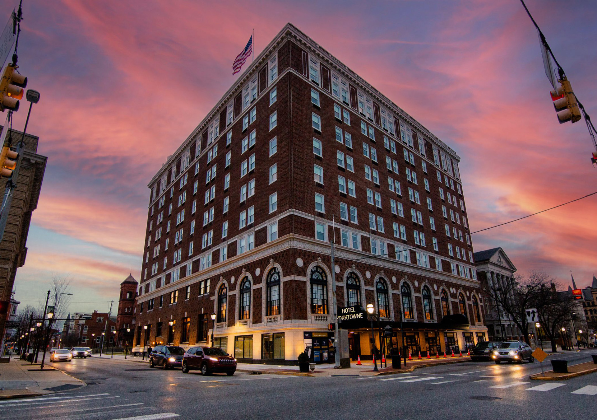 building-the-yorktowne-hotel-hero-image Tall brick building with a sign saying, "Hotel Yorktowne", in a downtown area with other brick buildings in the background. The sky is a pink and blue sunset.