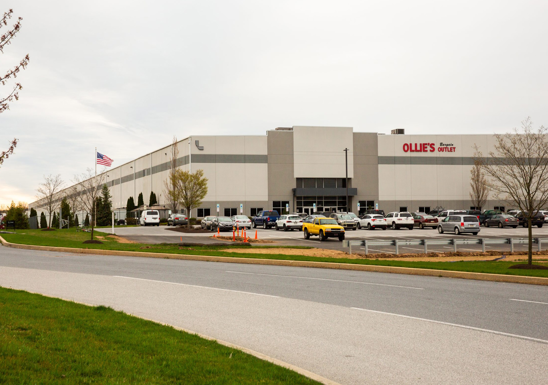 expanding-ollies-distribution-center-hero-image A tan and grey warehouse labeled "Ollie's Bargain Outlet," with numerous cars parked outside and an American flag displayed in the parking lot, all under an overcast sky.