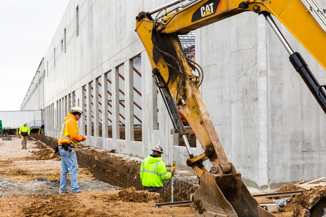 Massive light grey concrete walls that has rectangular openings for windows with steel framework supporting the building. There is a trench next to the concrete wall with men in high visibility vests and hardhats inside of if. In the foreground is a yellow excavator.
