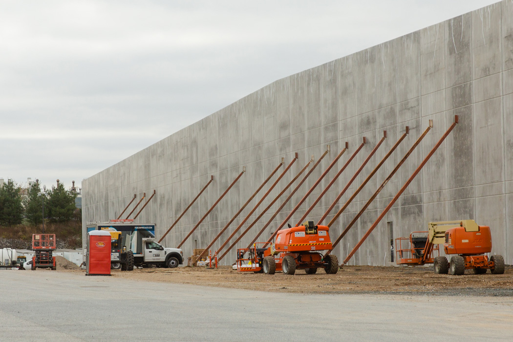 A large gray concrete wall with orange steel support beams stands alongside construction trucks, set against a backdrop of an overcast sky.