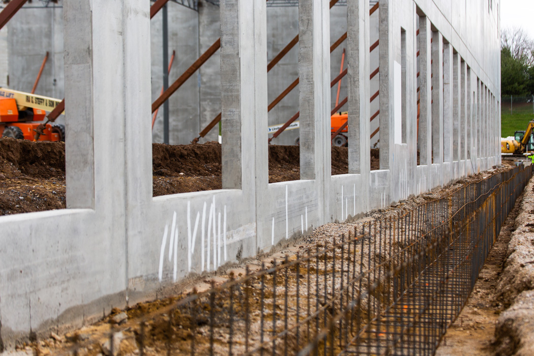 Massive light gray concrete walls with rectangular window openings are supported by a steel framework, with a trench running alongside the structure.