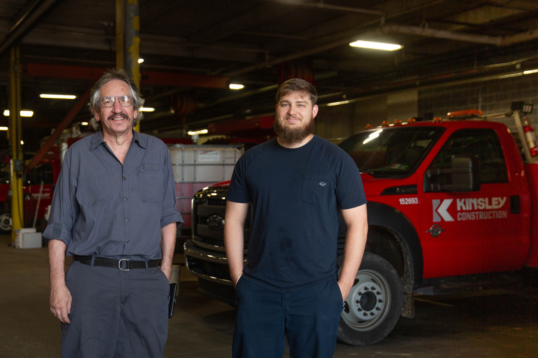 Two men in a body shop with red trucks and equipment in the background