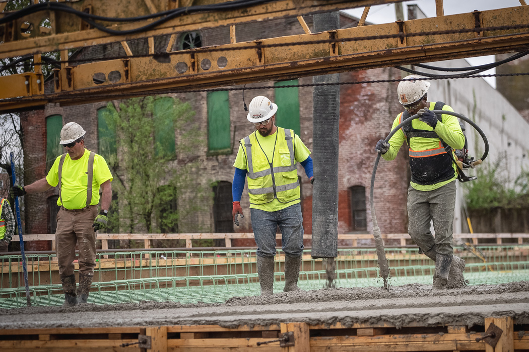 Three construction workers are focused on concrete work in an urban setting, with one using a concrete pump and the others supporting. The scene features an older brick building, a concrete slab, and machinery for pouring.