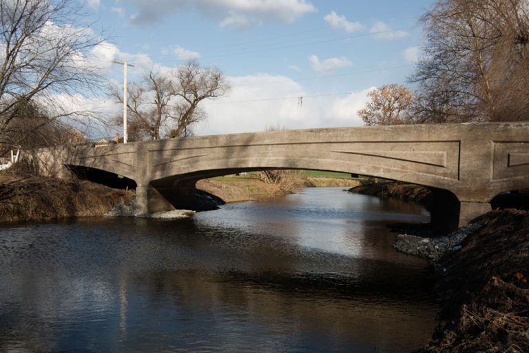A concrete bridge spans a river, framed by trees and set against a backdrop of cloudy blue skies.