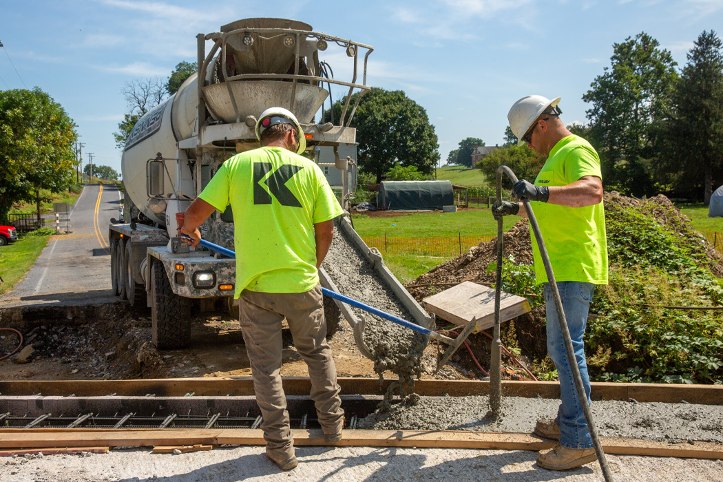 Two men in hard hats and high visibility shirts pouring concrete into a trench with a concrete mixer. Lush trees and blue skies are in the background.