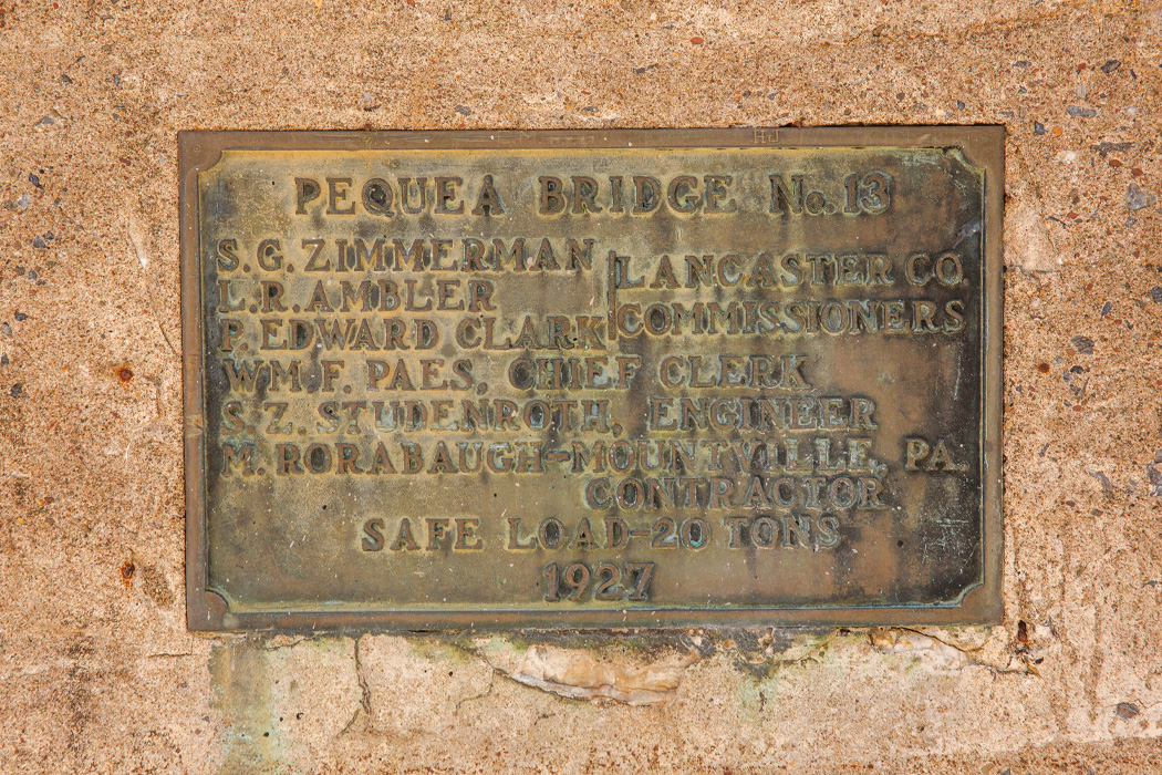 An old copper plaque titled "Peque Bridge No. 13" lists the individuals involved in its construction, with "1927" engraved at the bottom. The plaque is affixed to a concrete wall.