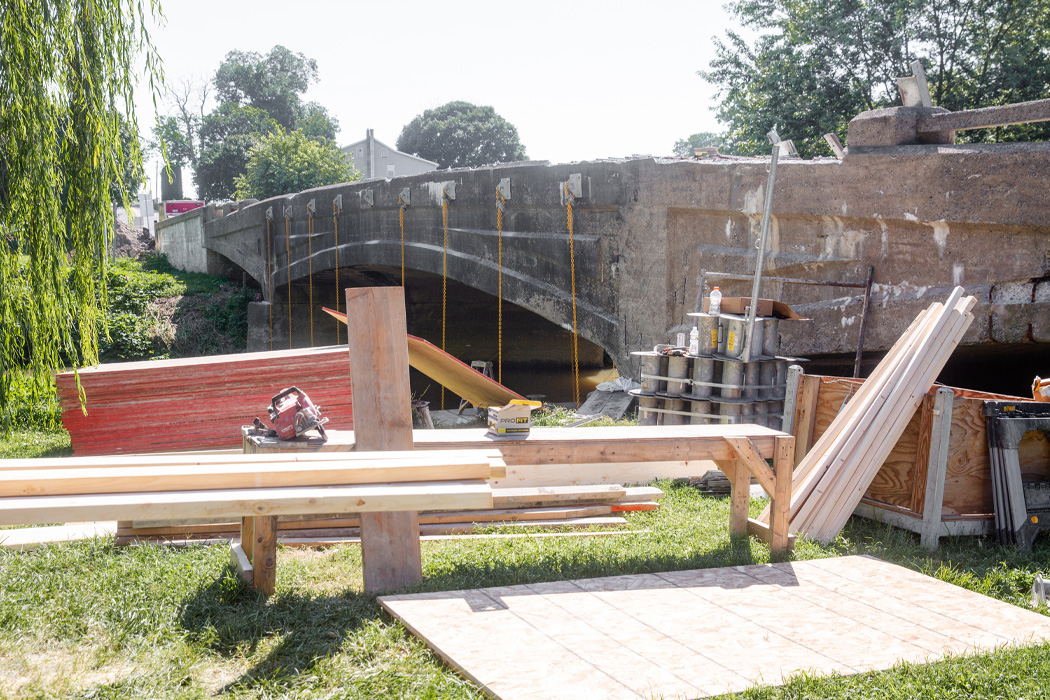 A concrete overpass spans a brown river, framed by green grass, lush trees, and overcast skies. In front of the overpass, wood beams and construction equipment are scattered across the site.