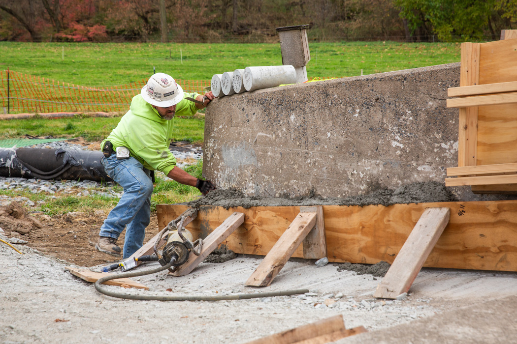 A construction worker in a green vest and hard hat smooths concrete on a large structure supported by wooden beams, standing on gravel with a grassy field in the background.