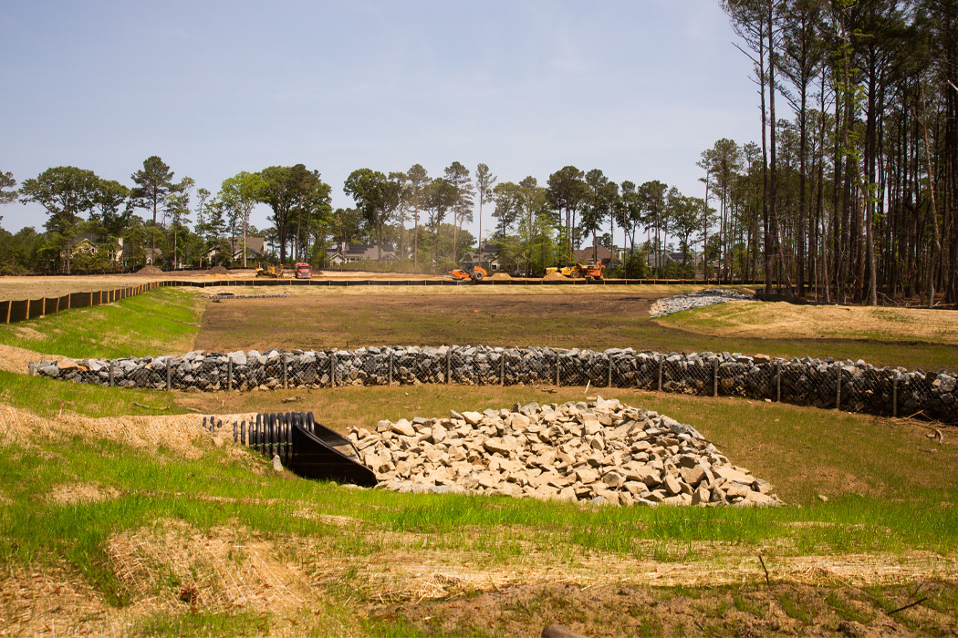 A expansive field with a large pile of rocks and orange construction equipment in the distance, set against a backdrop of trees and blue skies.
