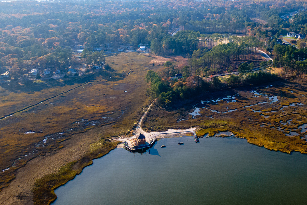 An aerial view of a gazebo perched on the banks of a lake, surrounded by a marsh and framed by a lush forest in the distance.