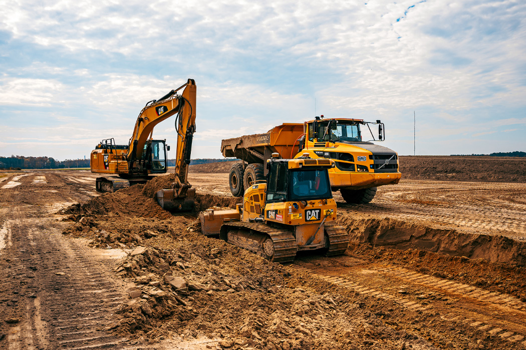 A yellow excavator, a yellow dozer, and a large yellow construction vehicle digging up dirt with cloudy skies in the background.