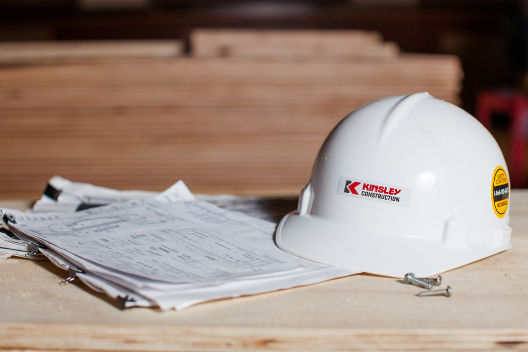 building-preconstruction-hard-hat-drawings A white hard hat with a Kinsley Construction logo rests on blueprints and plywood.