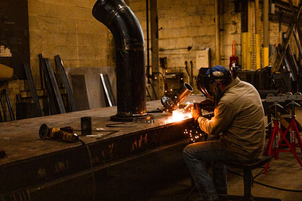 A welder works on a large metal structure in a dimly lit workshop.