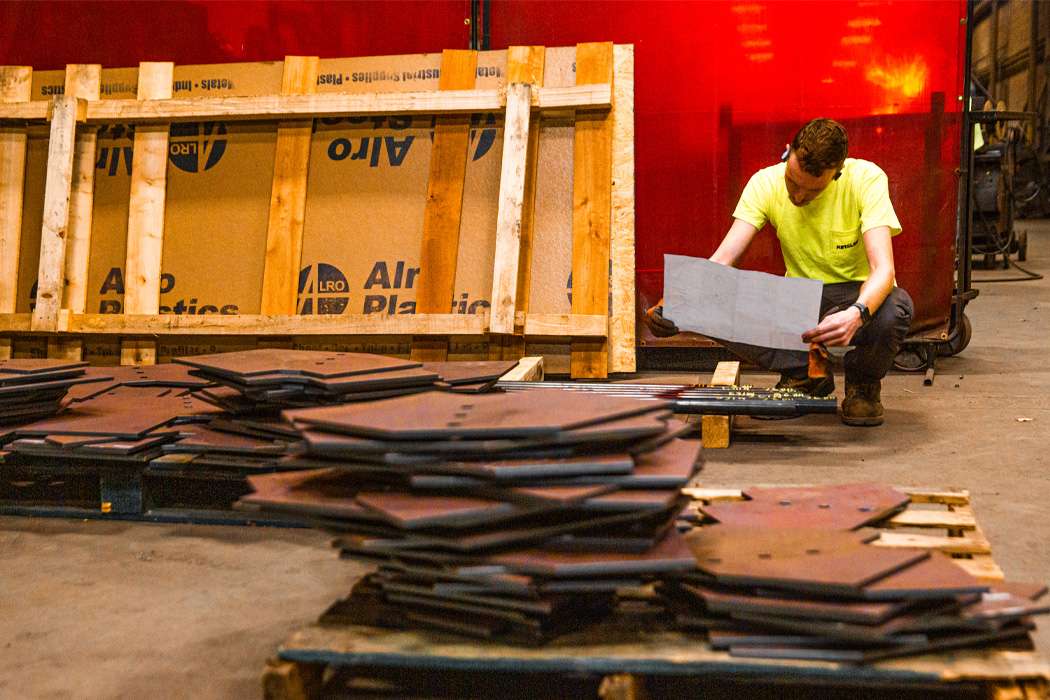 A worker examines a blueprint amidst stacks of metal parts in a factory.