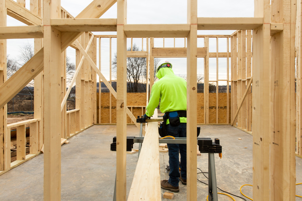 Man in a hard hat and high visibility sweatshirt inside of a wooden building frame sawing a wood beam.