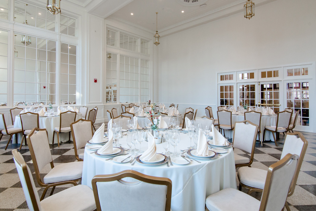 A dining area featuring a checkered floor, white upholstered chairs, and white circular tables with white place settings.