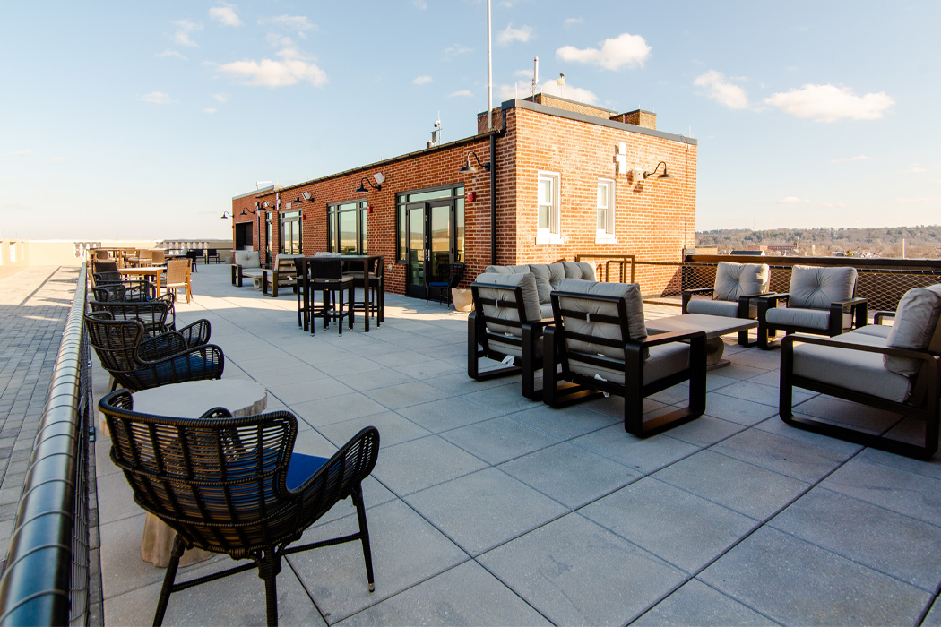 A rooftop patio featuring brown coffee tables and cushioned chairs, all set against a backdrop of the sky with some clouds.