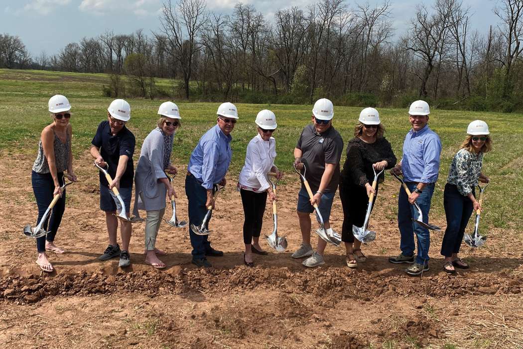 Nine people with hard hats and shovels digging into the ground at a groundbreaking event with grass and trees in the background.