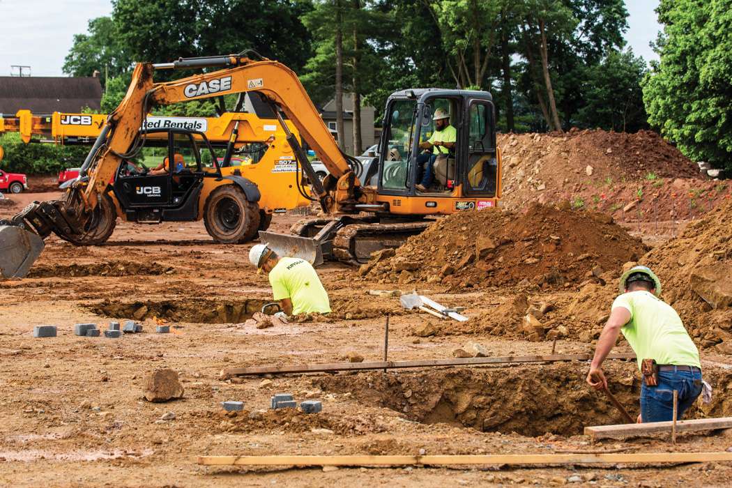 Men in hard hats and high visibility t-shirts digging holes with a yellow excavator and trees in the background