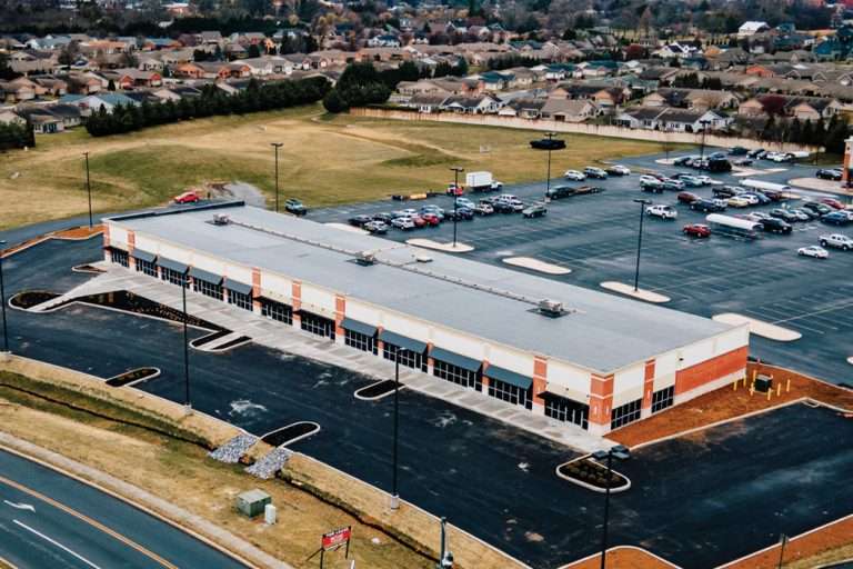 Large orange brick commercial building with empty storefronts. There is a large parking lot in the background filled with cars.