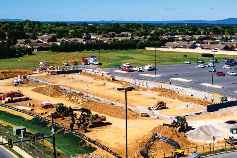 Heavy machinery works on a large construction site next to a nearly-empty parking lot.