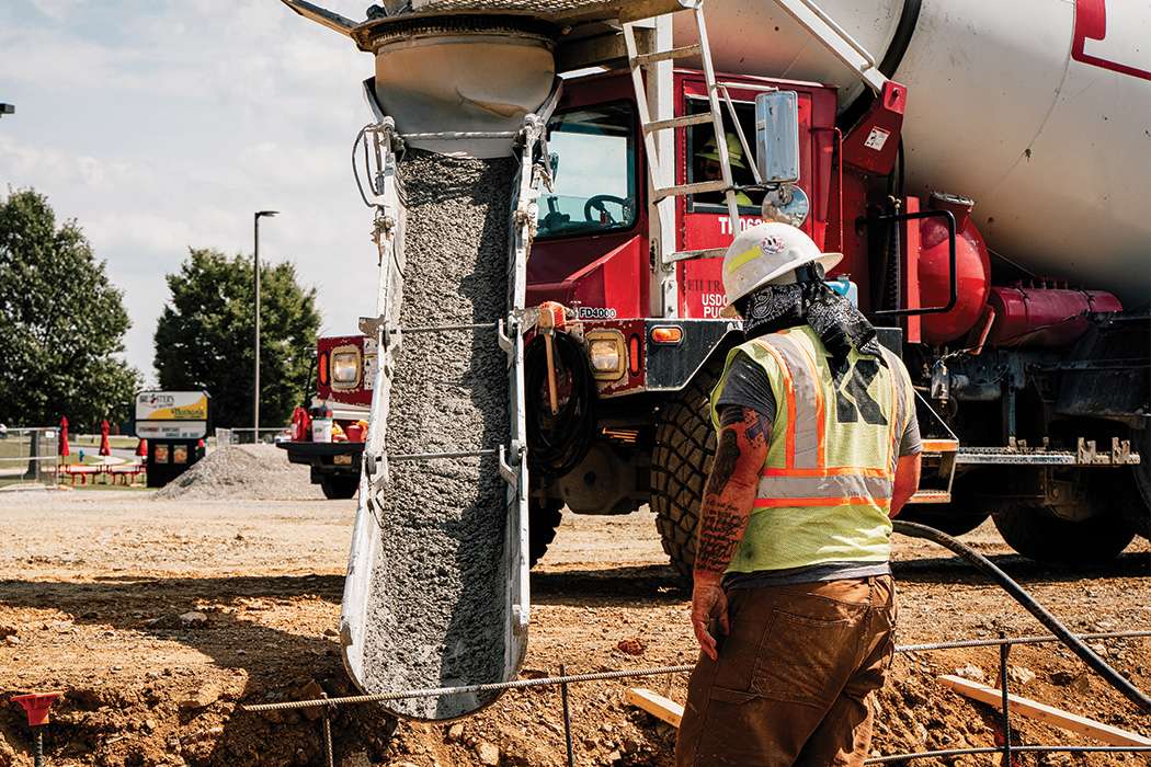 Man in high visibility vest and hard hat observing concrete being poured, from a concrete mixer, into a trench.