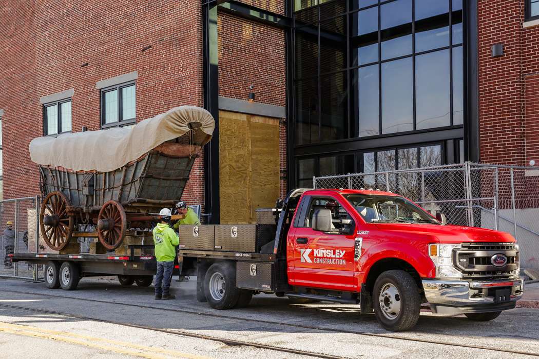 A red truck transports a covered wagon on a flatbed trailer in front of a brick building.
