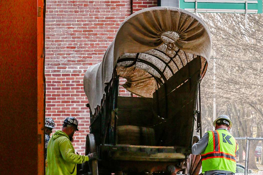 Men in high visibility jackets and hard hats bringing in a blue wooden Conestoga wagon