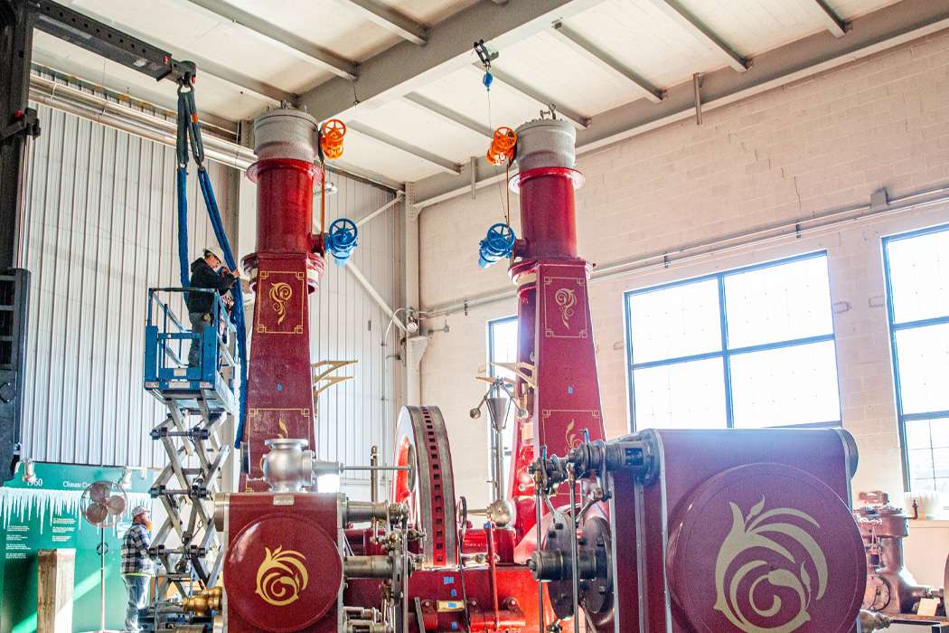 Workers install a large, ornate red steam engine in a spacious industrial building.