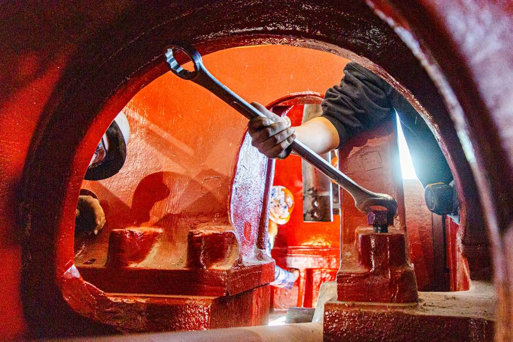 A worker uses a wrench to tighten a bolt inside a large, red piece of machinery.