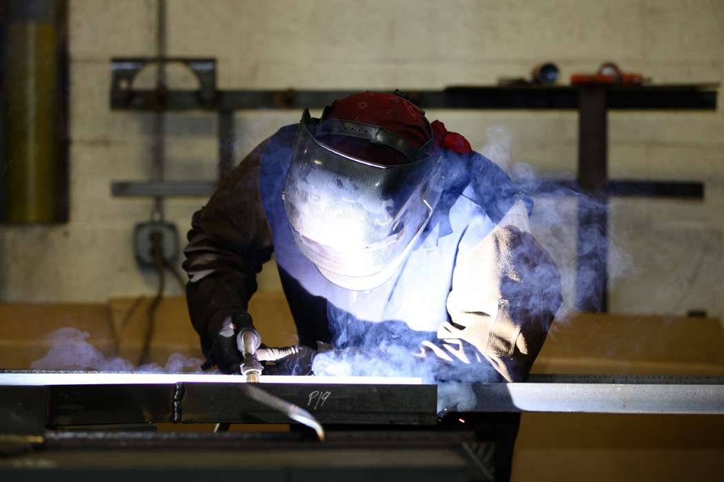 A welder wearing protective clothing works on a metal beam, surrounded by smoke and sparks.