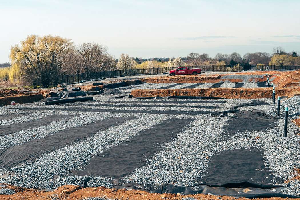 Construction site with gravel, black landscaping fabric, drainage pipes, a red pickup truck, and trees, focusing on ground preparation for future development.