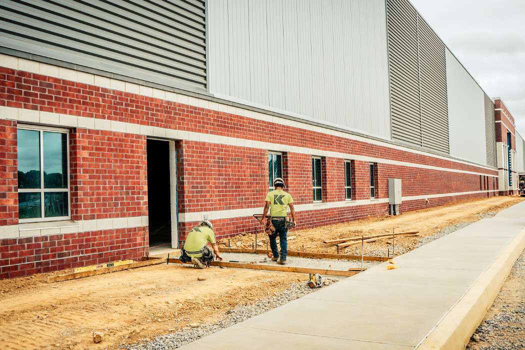 Two construction workers wearing hard hats and neon shirts are pouring concrete at the base of a new brick building.