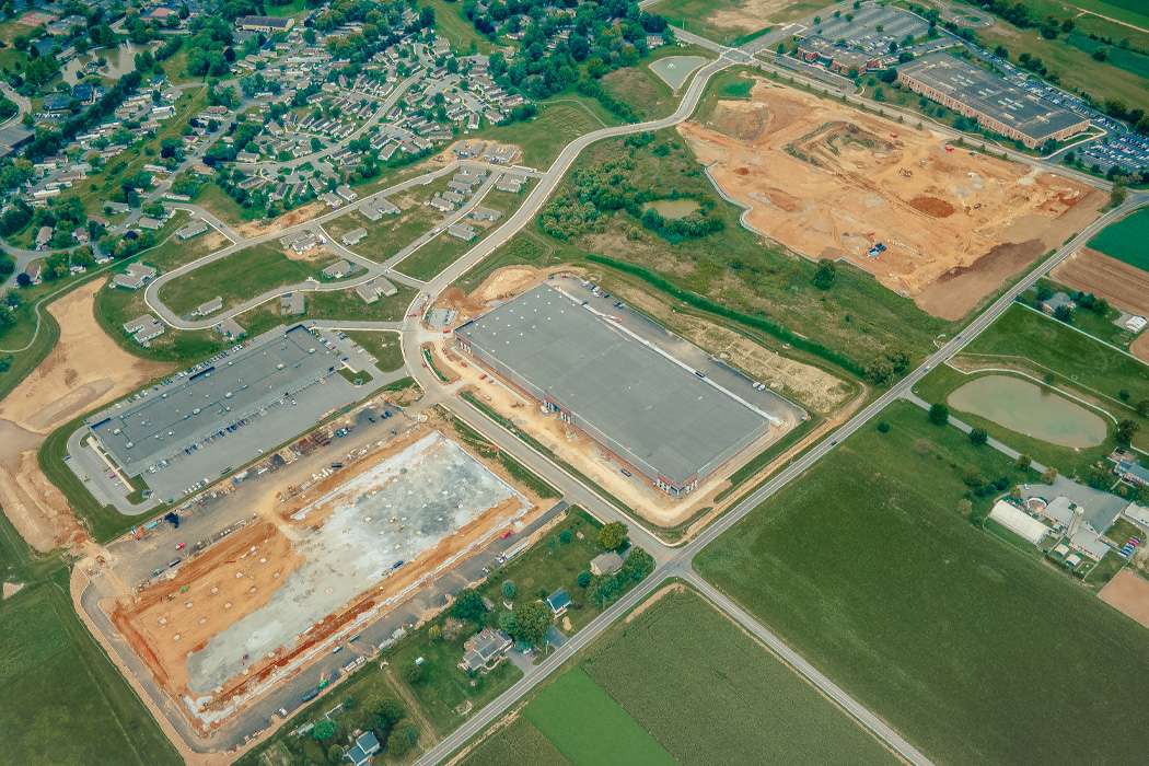 An aerial view captures a sprawling suburban landscape with a new warehouse surrounded by a paved parking lot, adjacent construction, a dense residential neighborhood, and a field with a pond.