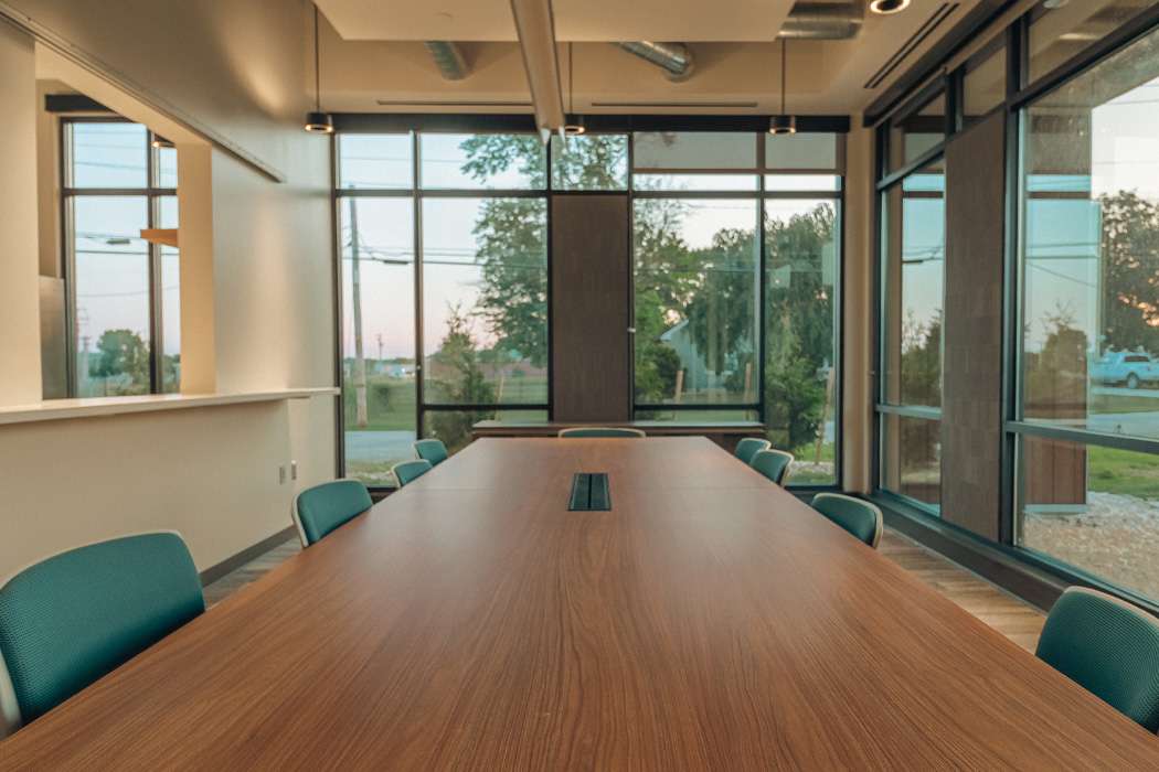 A long conference table sits in a modern room with large windows overlooking a tree-lined landscape.