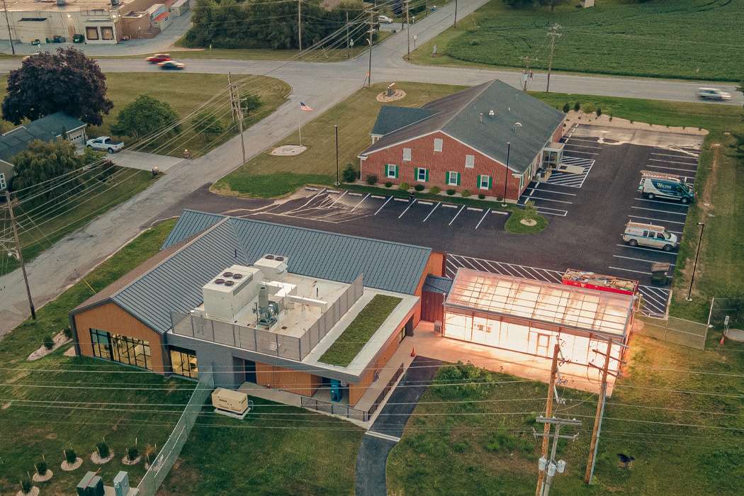 An aerial view shows a modern building complex, including a greenhouse, next to a brick building in a rural setting.