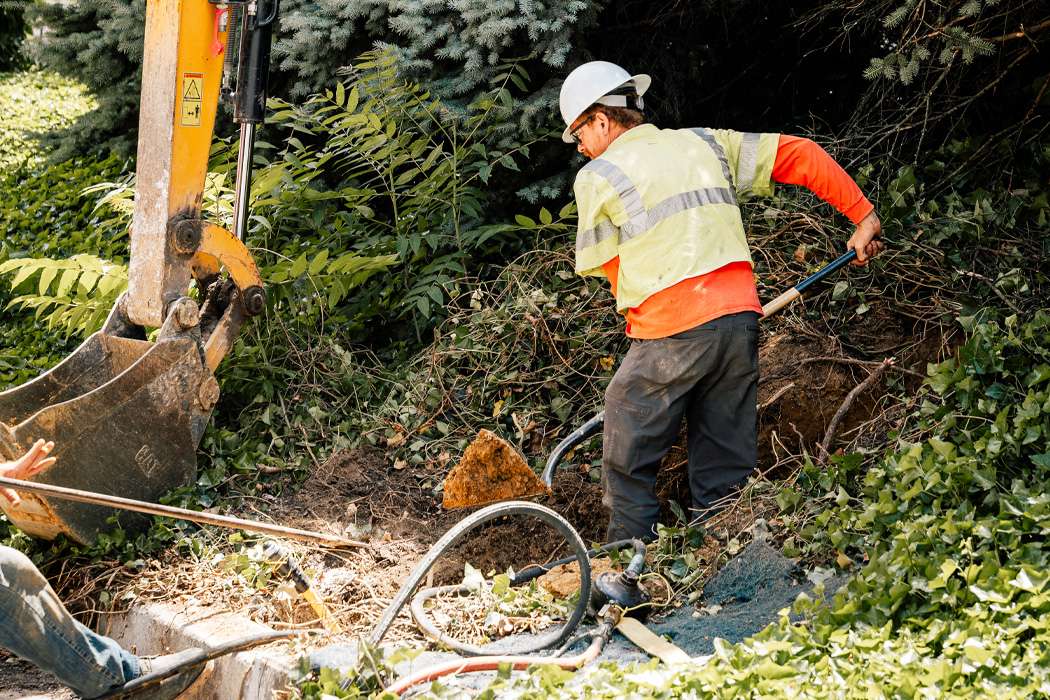A worker uses a shovel and excavator to clear brush near a utility line.