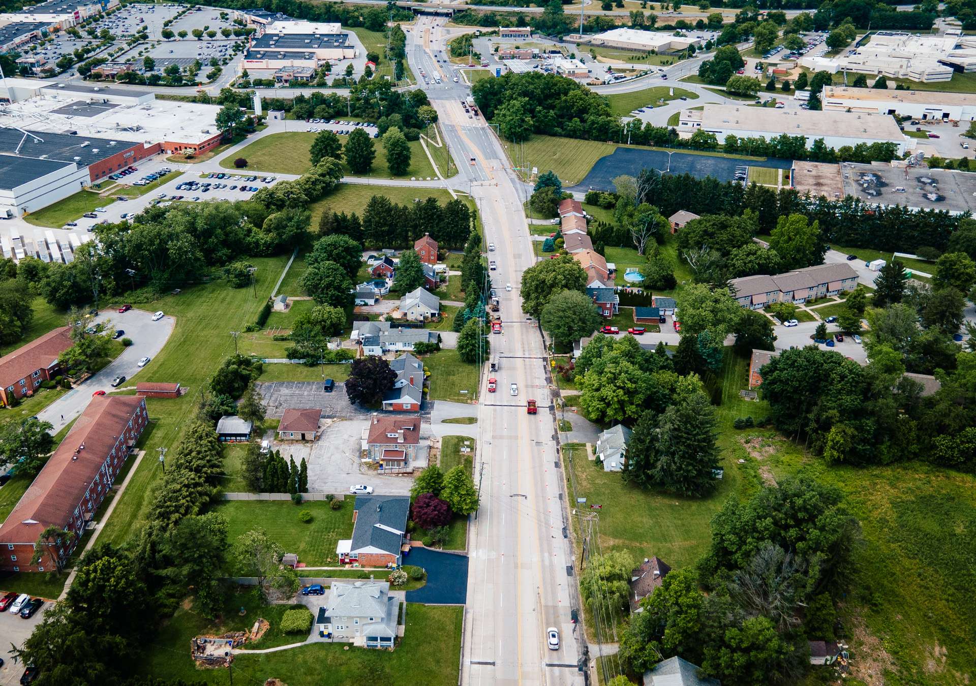 mt-zion-gas-main-replacement-hero Aerial view of a suburban neighborhood with a straight road lined by trees, houses, and businesses, including a shopping center. The area is surrounded by a grassy field, under a clear blue sky.