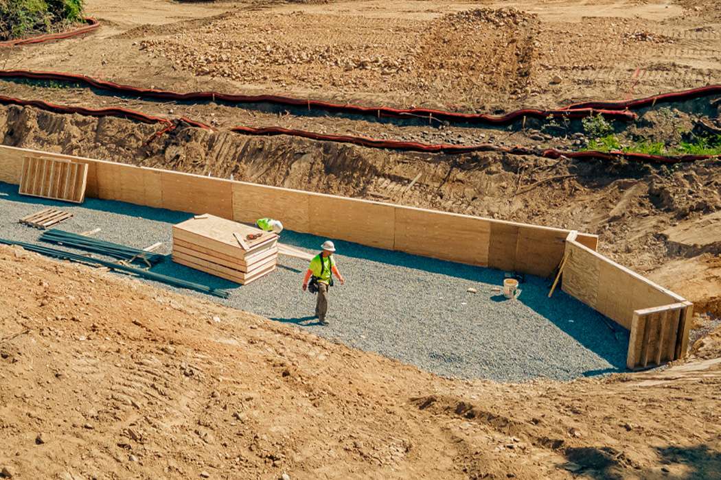 A construction worker in a high visibility shirt walking on a gravel path beside a plywood wall under construction on a sloped embankment, with materials scattered around.