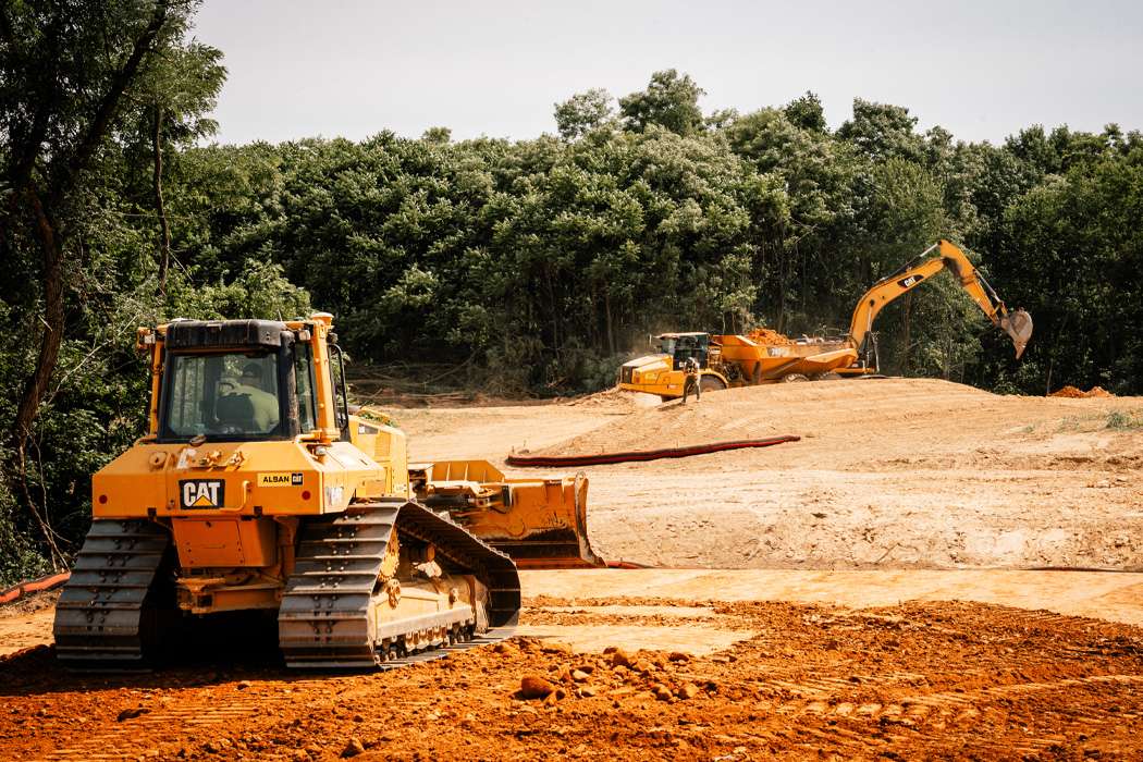 A yellow dozer and yellow excavator digging large piles of dirt with green trees in the distance