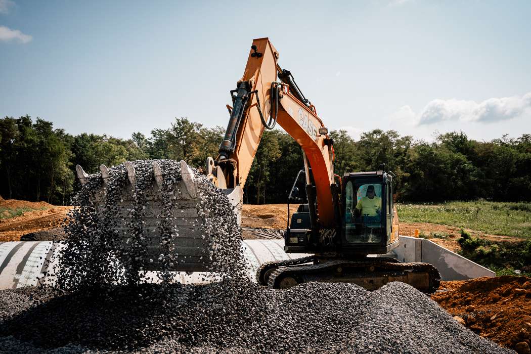 Yellow excavator picking up a large pile of grey rocks with a lush forest and cloudy skies in the background.