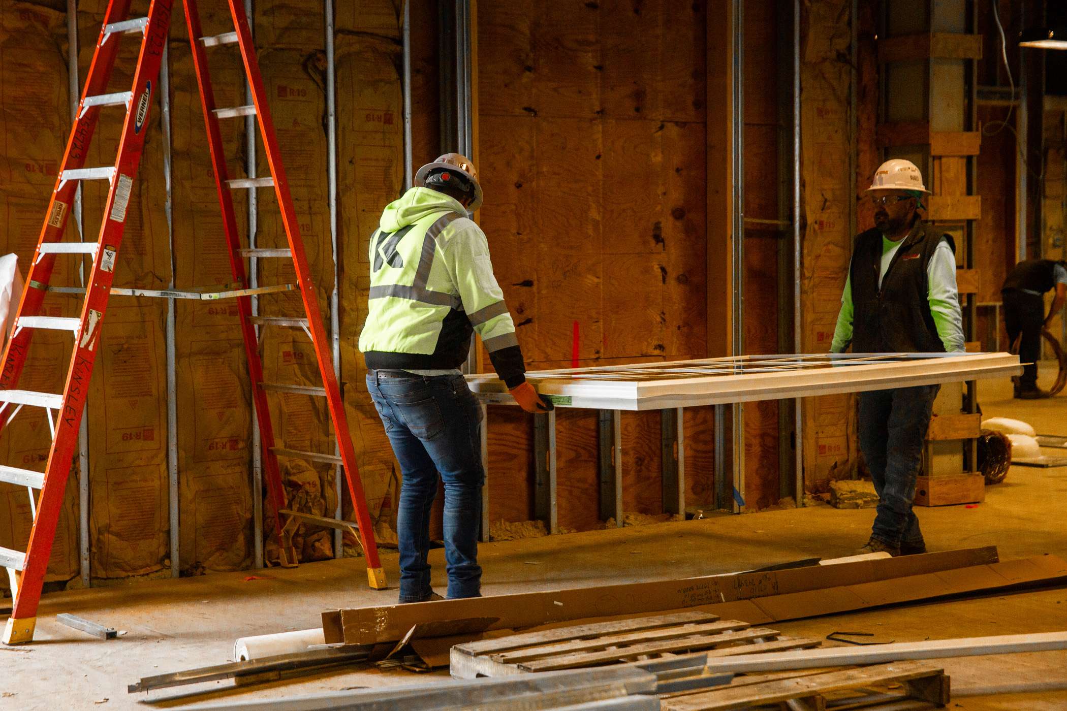 Two construction workers maneuver a piece of lumber inside a building under construction.