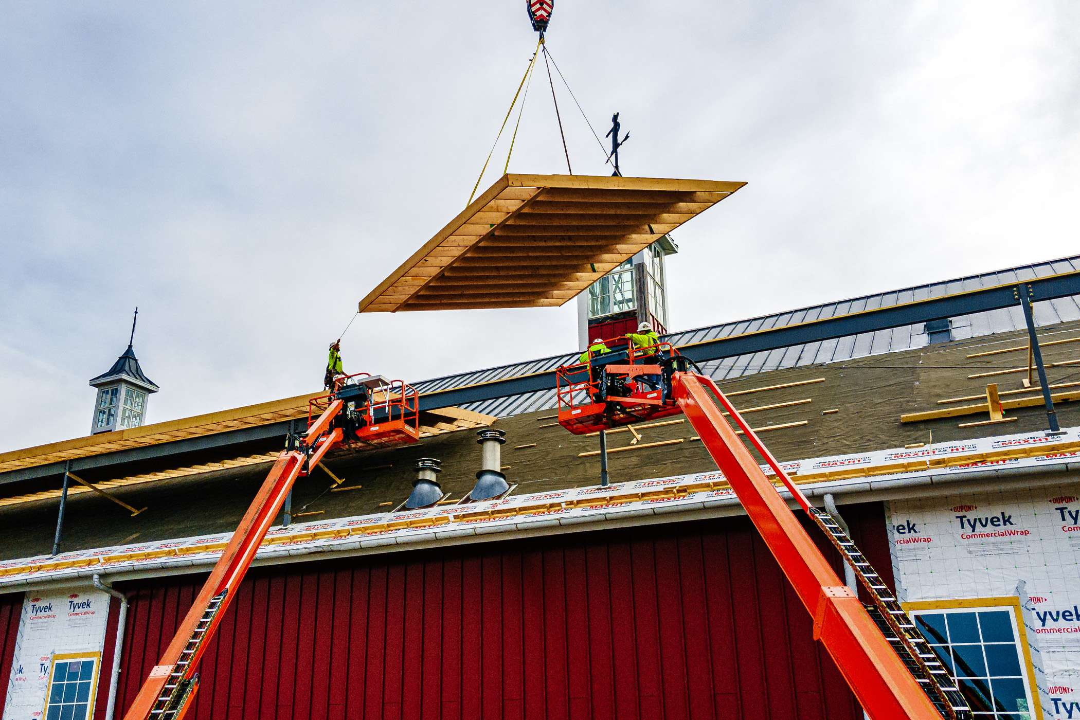A crane lifts a large wooden panel onto a building's roof using a pair of extending arms.