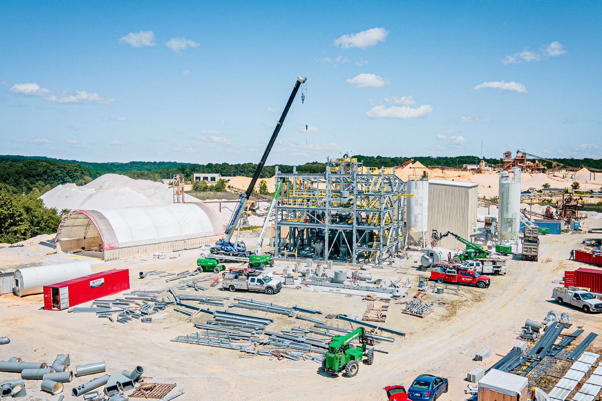 A view of a large steel building being built with a crane. There are blue skies and green trees in the background.