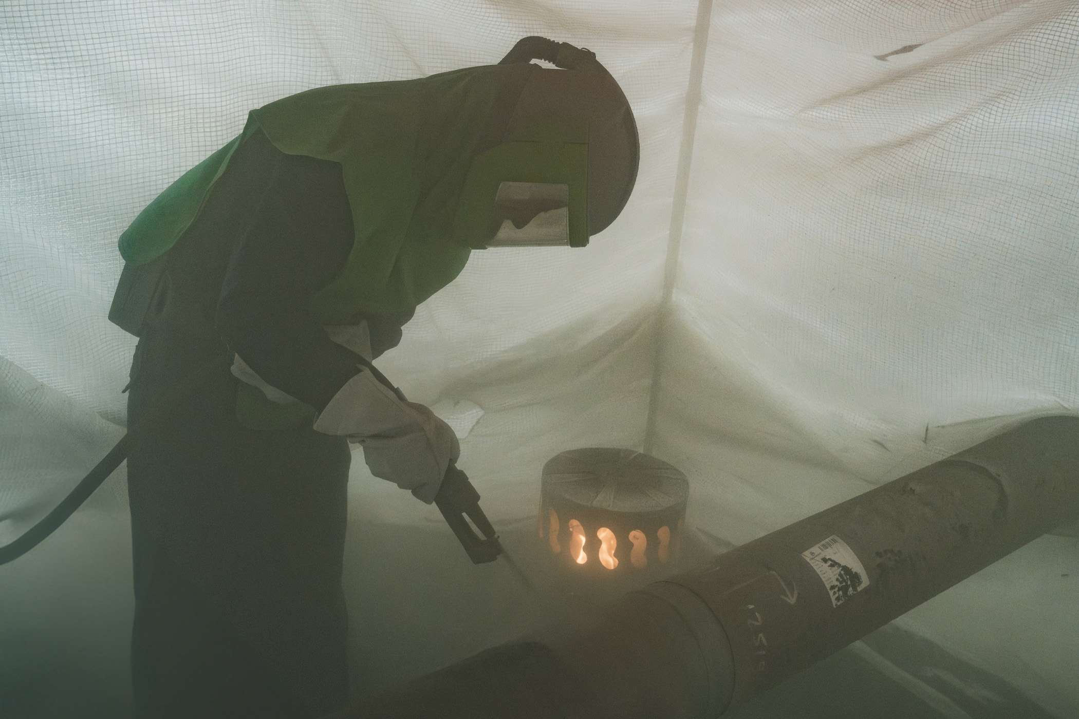 Man in protective gear is welding a pipe with a torch in a tent.
