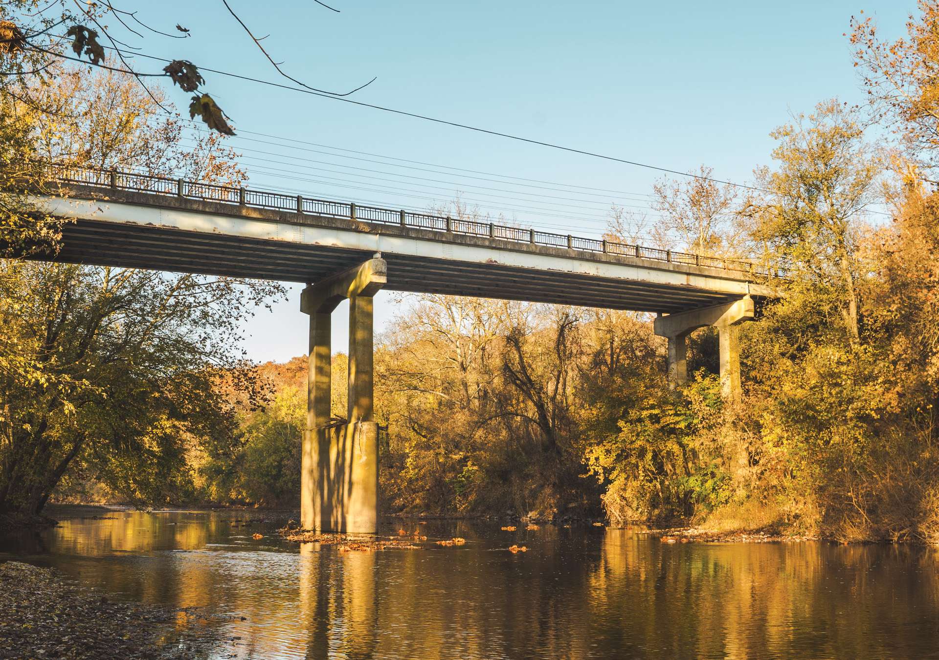 conewago-creek-and-bridge-hero-image A bridge spans a calm river bordered by autumnal trees.