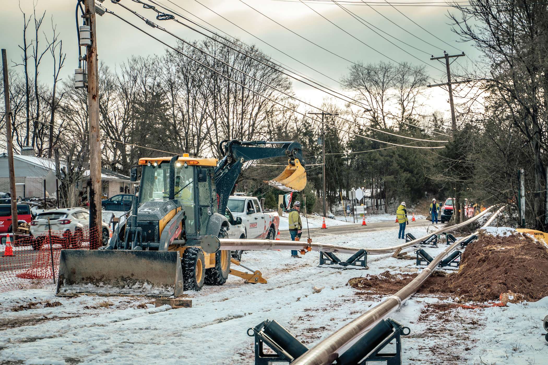 Workers install a pipeline in a snowy residential area using heavy machinery.