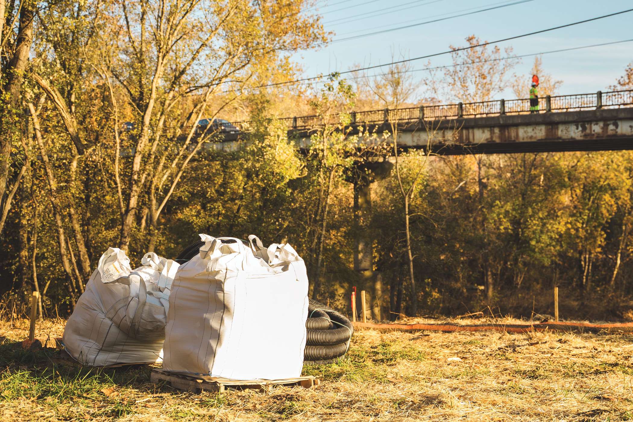 Large white bags sit near a bridge in a rural autumnal setting.