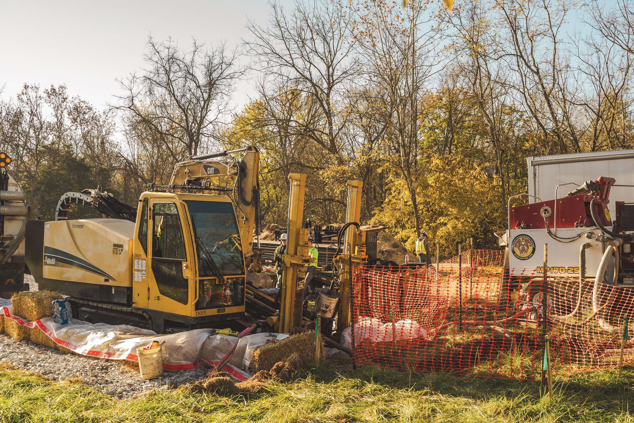 Two pieces of heavy machinery sit on a construction site surrounded by a temporary fence and autumn foliage.