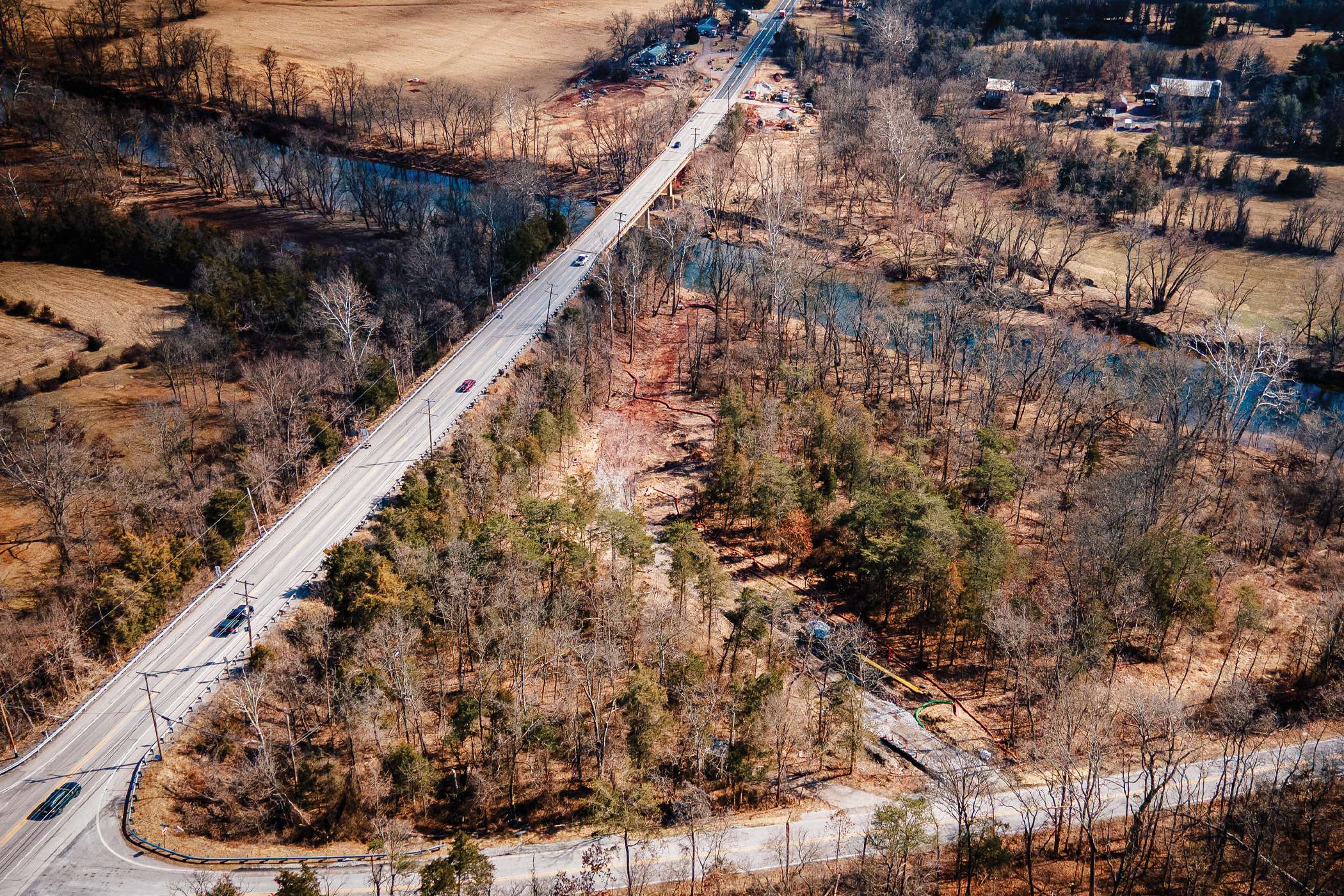 An aerial view shows a highway bridge crossing a river, surrounded by winter woods.